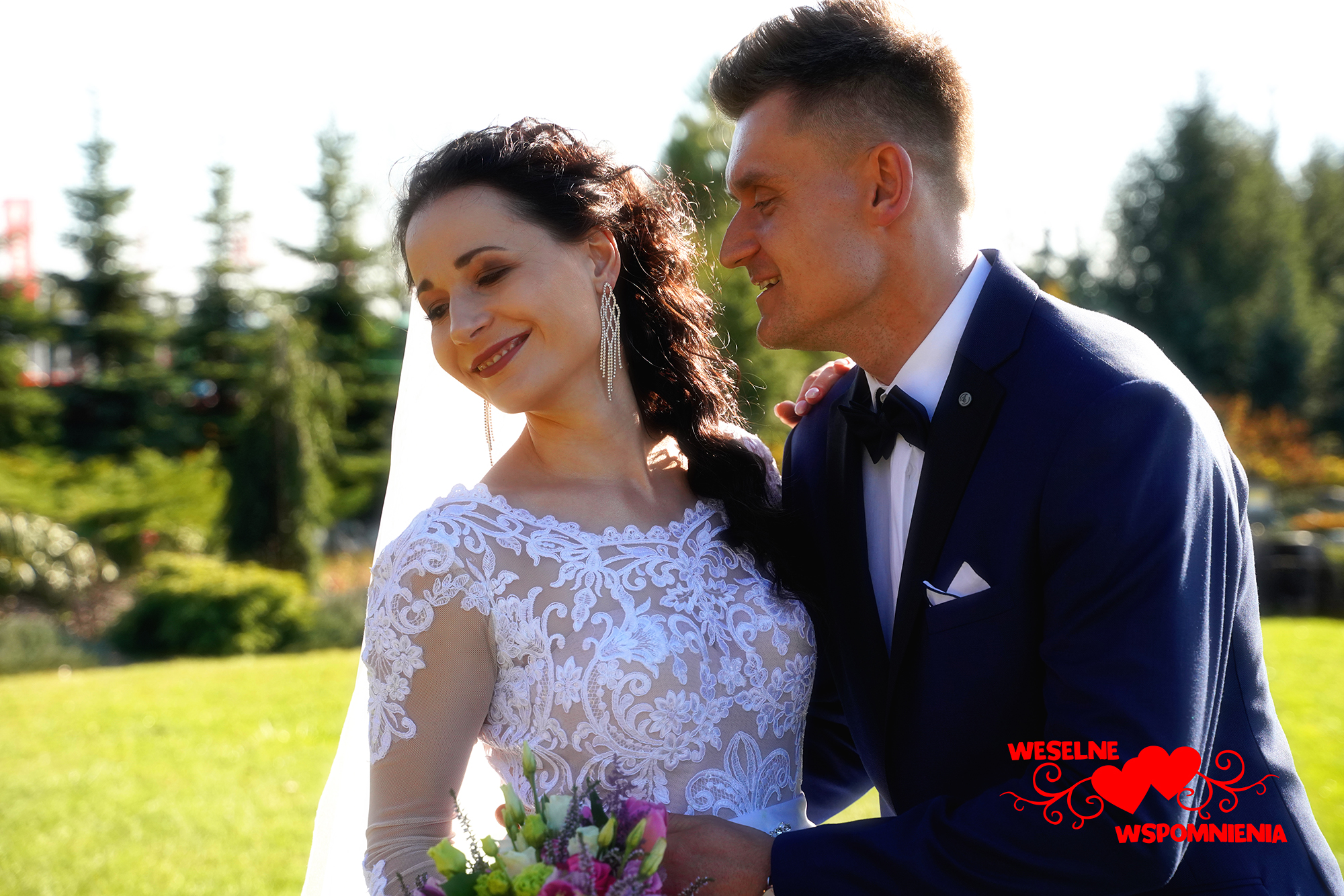 Uśmiechnięta panna młoda w koronkowej sukni i długich kolczykach spogląda w dół, obok niej pan młody w granatowym garniturze z muchą, w tle zielony trawnik i drzewa.