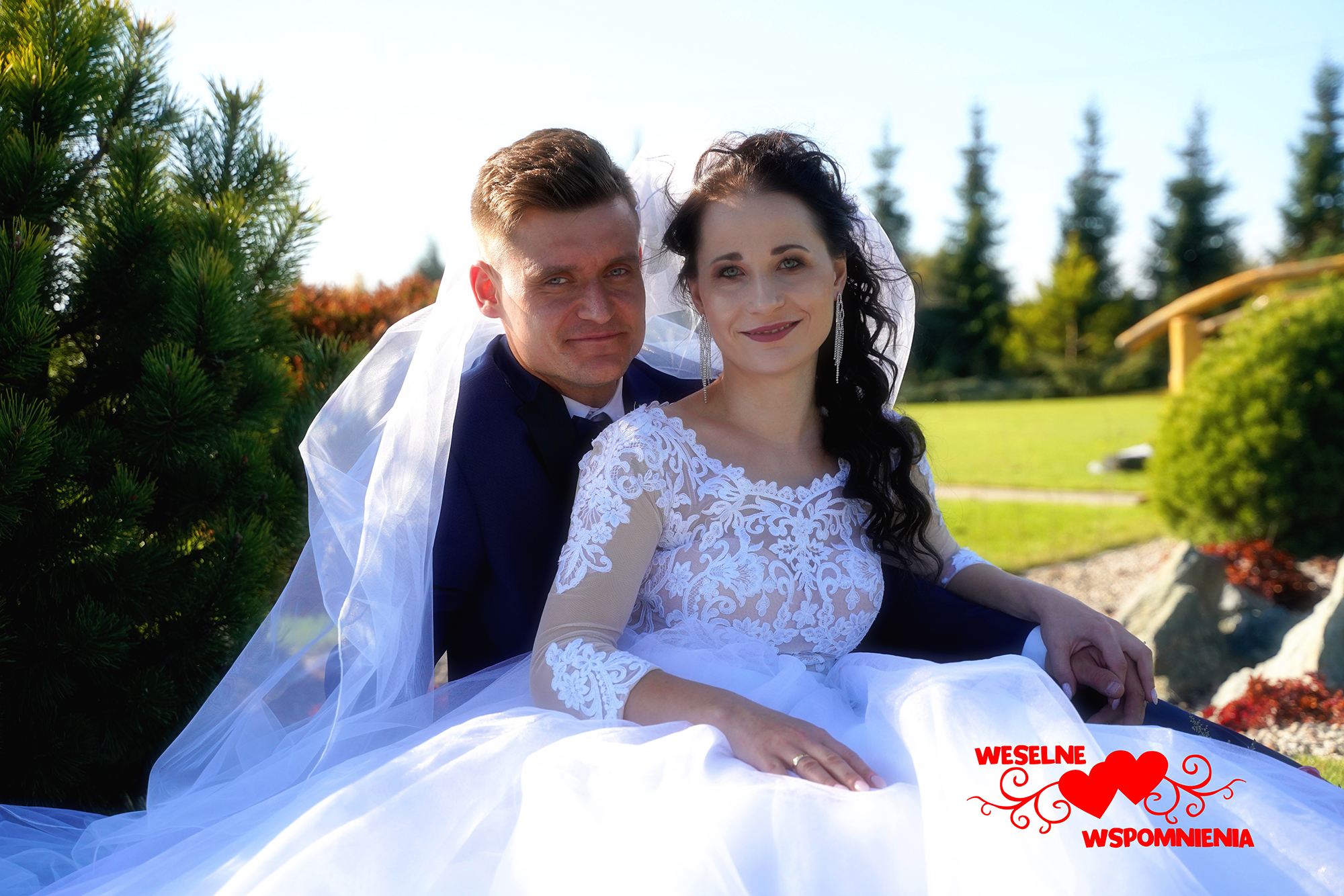 Para młoda, panna młoda w koronkowej sukni z długim rękawem, pan młody w granatowym garniturze, siedząca na trawie w plenerze, widoczny mostek w tle, logo 'Weselne Wspomnienia' w prawym dolnym rogu.