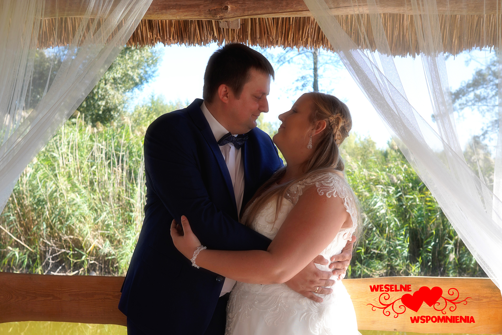Para młoda w objęciach pod drewnianą konstrukcją z białymi zasłonami, widoczne trzciny i drzewa w tle, na pierwszym planie naklejka z napisem 'Weselne Wspomnienia' i czerwonymi sercami.