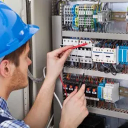 Mężczyzna w niebieskim kasku, w koszuli w kratę, dokonuje pomiarów w rozdzielnicy elektrycznej za pomocą miernika cęgowego z czerwonymi przewodami.