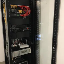 Czarna szafa rack z otwartymi drzwiami z przezroczystego szkła, wypełniona kablami i urządzeniami sieciowymi, takimi jak routery i switche, z żółtymi i czerwonymi kablami patchcord.