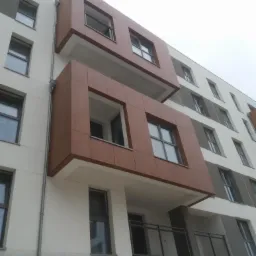 Fasada budynku z balkonami wykończonymi panelami HPL imitującymi drewno, widok z dołu, w tle pochmurne niebo.