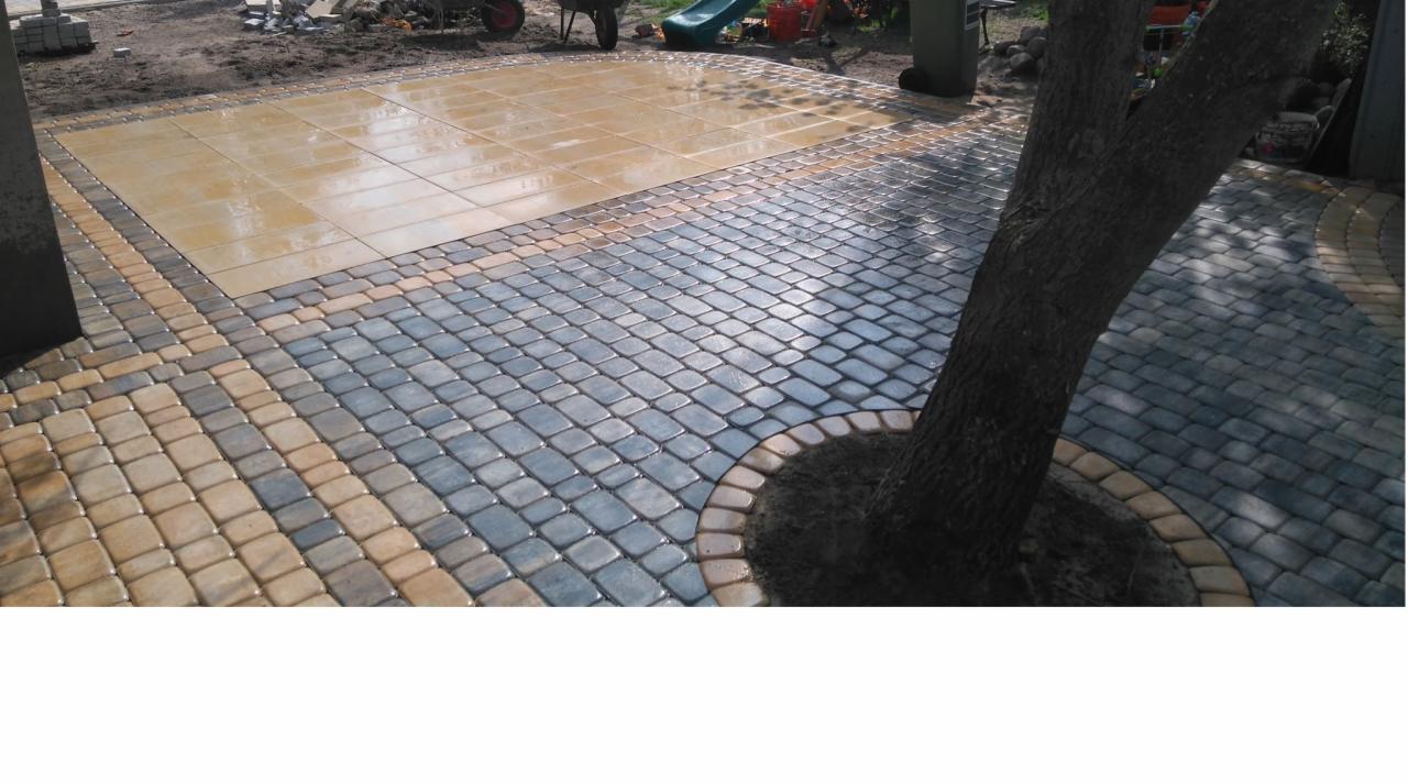 Ułożona nawierzchnia z kostki brukowej w dwóch odcieniach szarości i beżu, obramowana jaśniejszym odcieniem wokół pnia drzewa, z fragmentem większych płyt w tle, na częściowo przygotowanym podłożu.