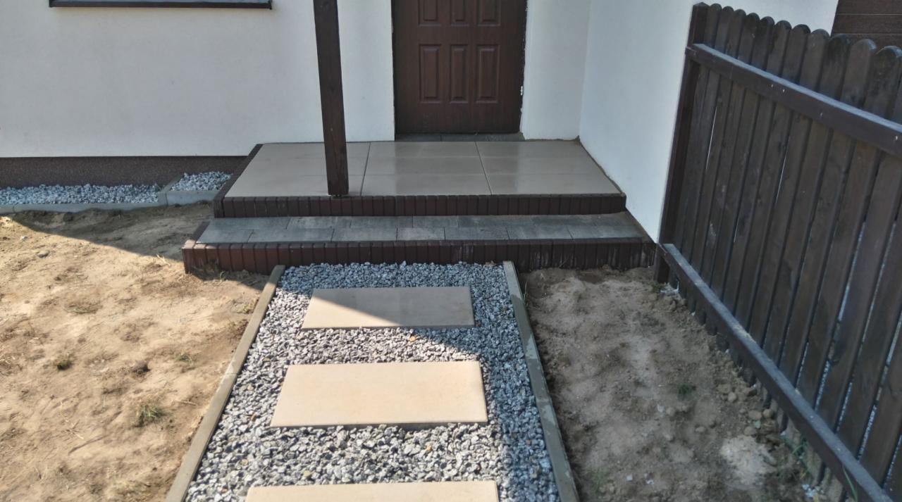 Wykończone wejście do domu z jasnymi płytami na schodach i ścieżką z płyt otoczoną drobnym żwirem, obok drewniane ogrodzenie.