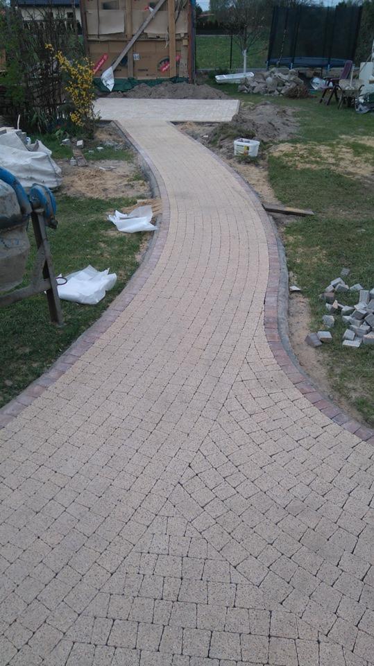 Zakrzywiona ścieżka z jasnej kostki brukowej z ciemniejszym obramowaniem, w tle widoczny częściowo plac budowy i betoniarka.