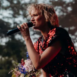 Wokalistka w cekinowej sukience śpiewa do mikrofonu na tle rozmytych drzew i bukietu kwiatów.