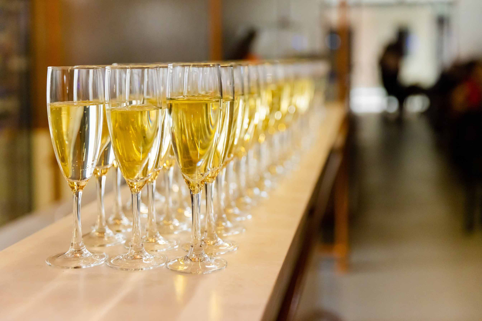 Rząd kieliszków do szampana wypełnionych musującym, złocistym napojem, ustawionych na jasnym blacie, z rozmytym tłem sugerującym wnętrze sali.