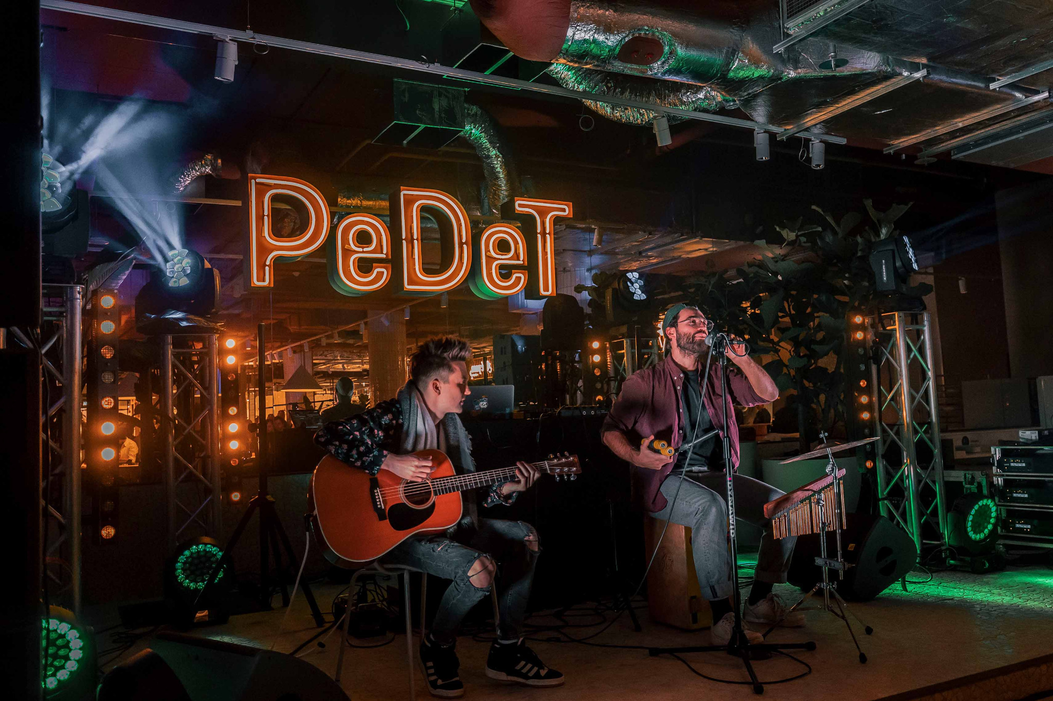 Duet muzyczny na scenie z gitarą akustyczną i instrumentami perkusyjnymi, podświetlony kolorowymi światłami, z neonowym napisem 'PeDeT' w tle.