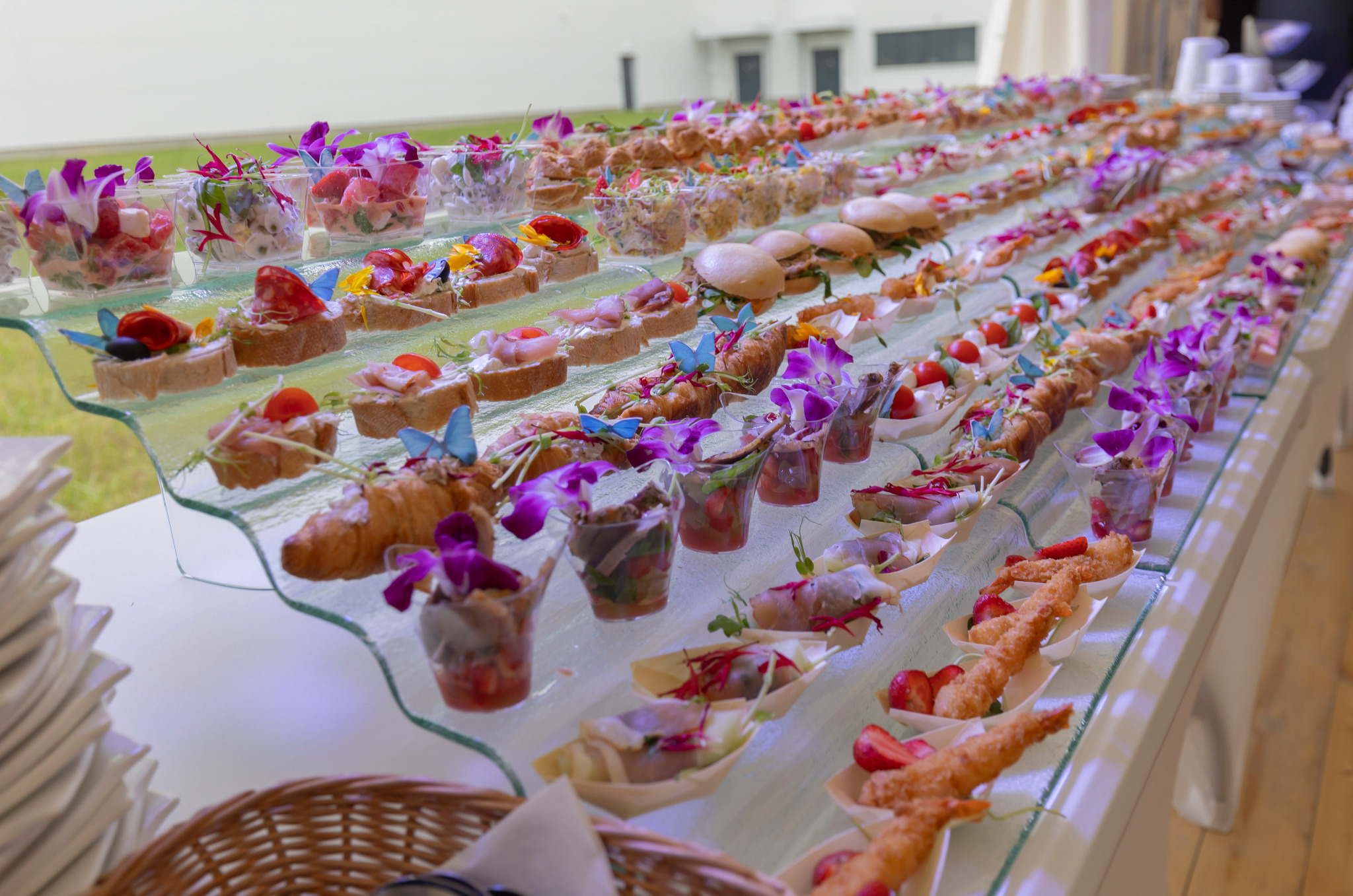 Szwedzki stół z różnorodnymi przekąskami: mini burgery, kanapeczki, sałatki w szklankach, krewetki w tempurze, wszystko udekorowane kwiatami i motylami, ustawione na szklanym podeście.