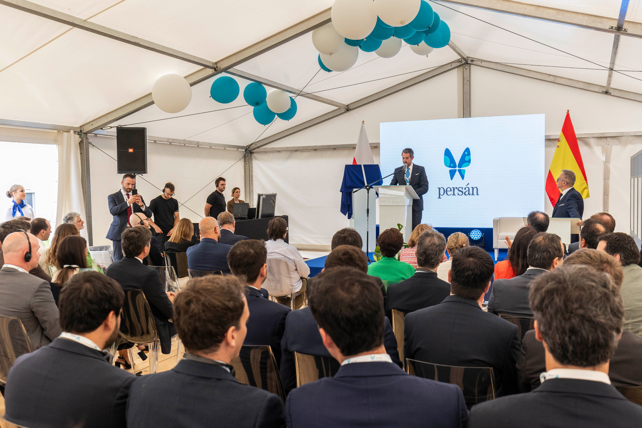 Konferencja w namiocie: prelegent przemawia zza mównicy przed zgromadzonym tłumem, w tle ekran z logo firmy, flagi, balony i obsługa techniczna.