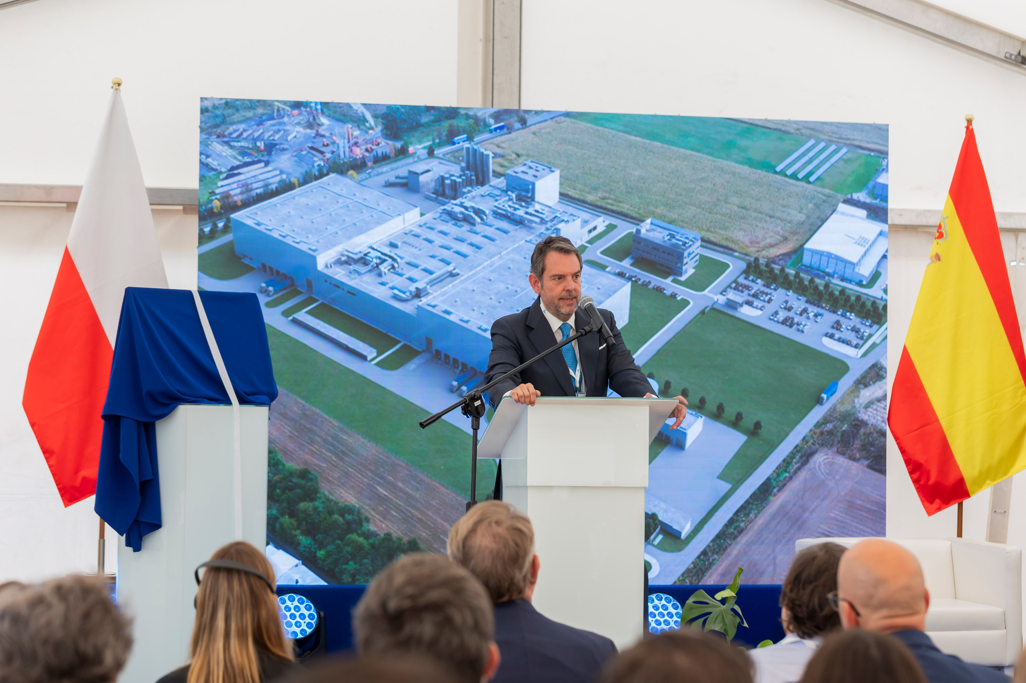 Prezentacja nowego zakładu produkcyjnego: mężczyzna w garniturze przemawia z mównicy na tle ekranu z widokiem lotniczym fabryki, z flagami Polski i Hiszpanii po bokach.