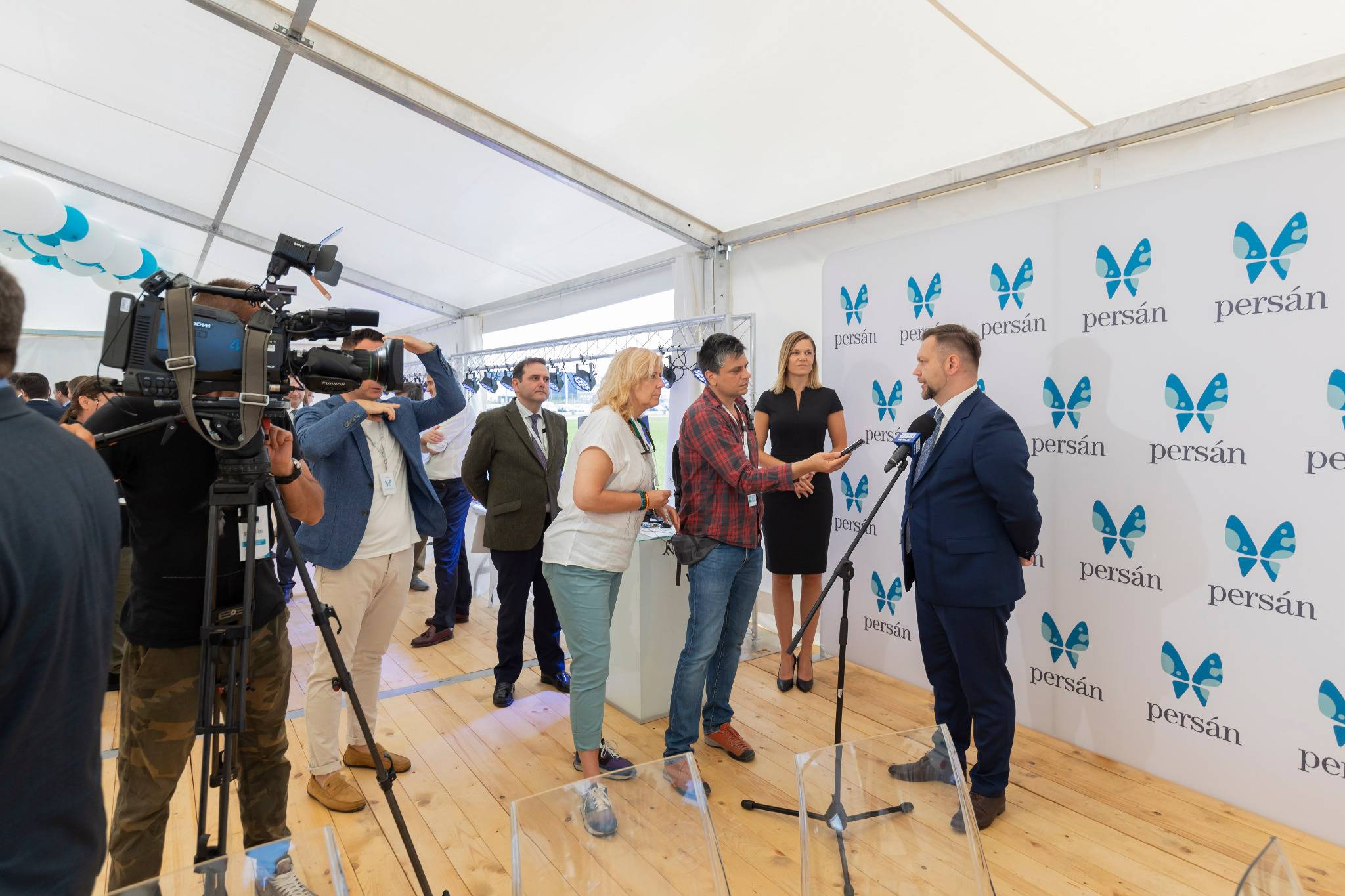 Wywiad z mężczyzną w garniturze na tle ściany z logo firmy 'persán', nagrywany przez ekipę filmową z profesjonalnym sprzętem, w przestrzeni eventowej pod namiotem.