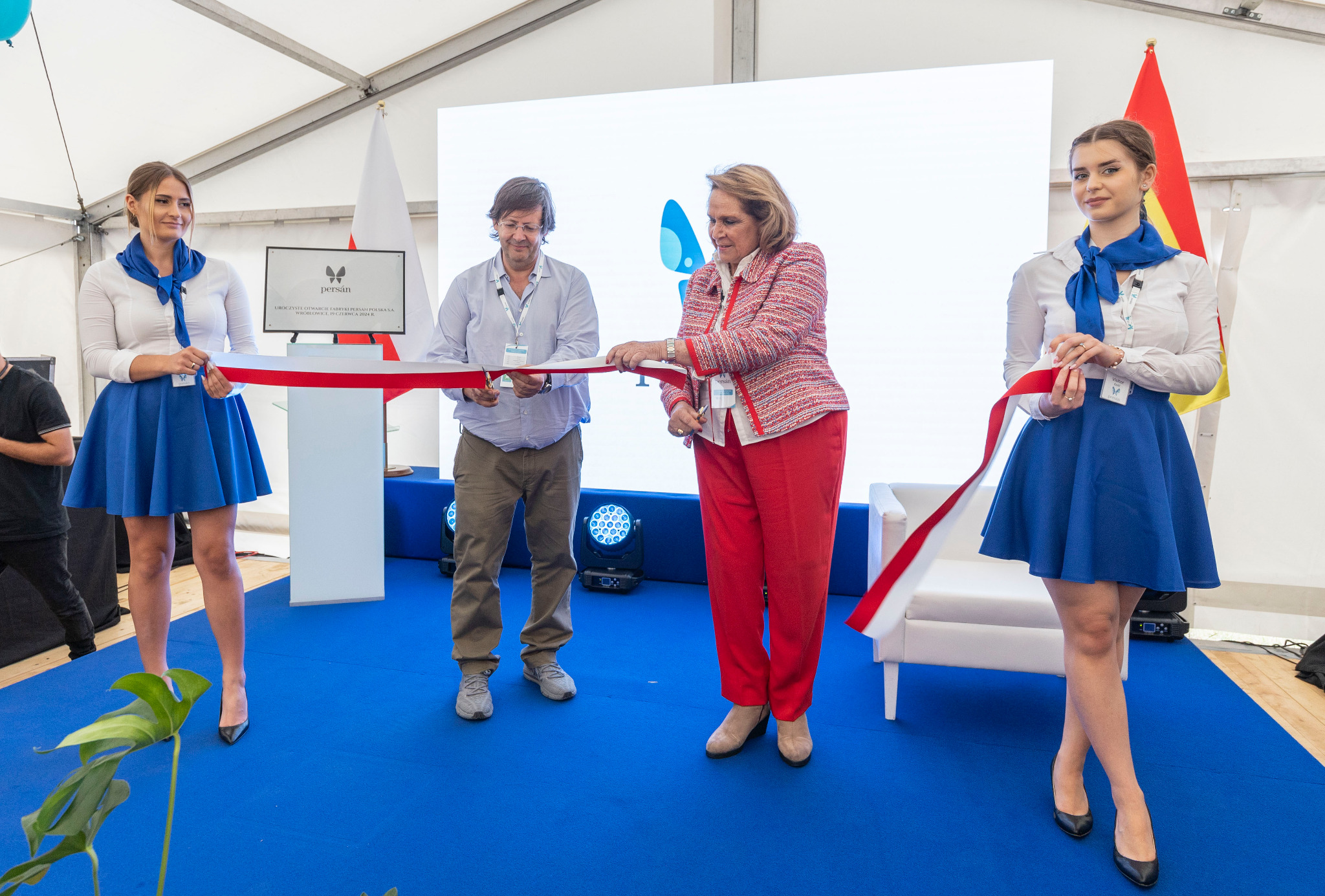 Uroczyste przecięcie wstęgi podczas otwarcia firmy; dwie kobiety w niebieskich spódnicach asystują, a dwoje gości przecina czerwoną wstęgę na tle logo firmy i flagi Polski i flagi innego kraju.
