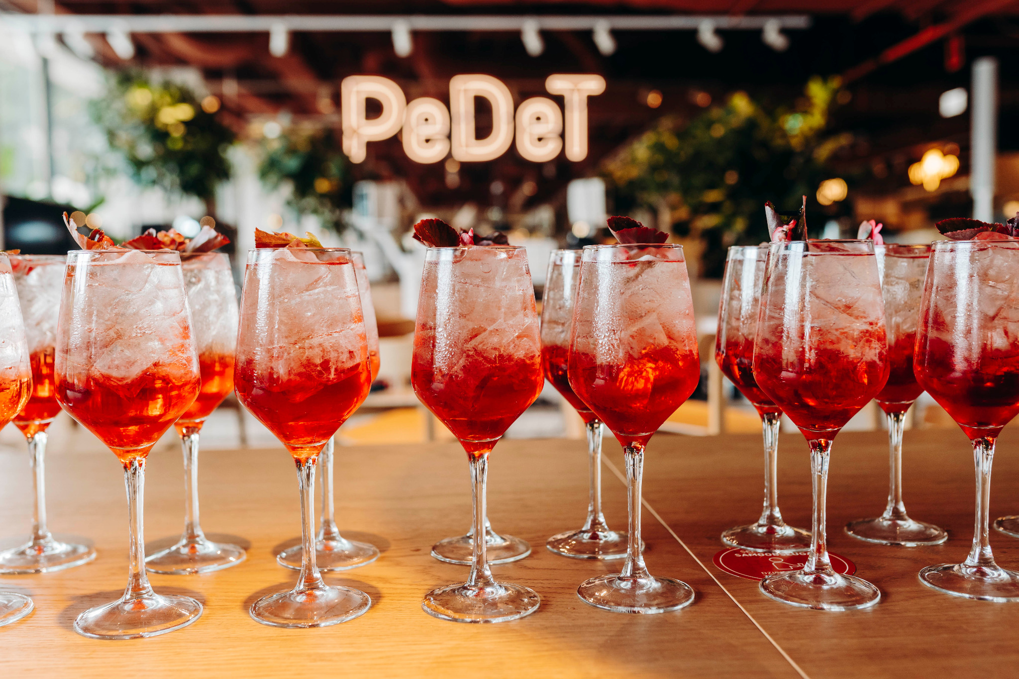 Rząd kieliszków z Aperol Spritz udekorowanych plasterkiem pomarańczy na drewnianym blacie, w tle neonowy napis 'PeDeT'