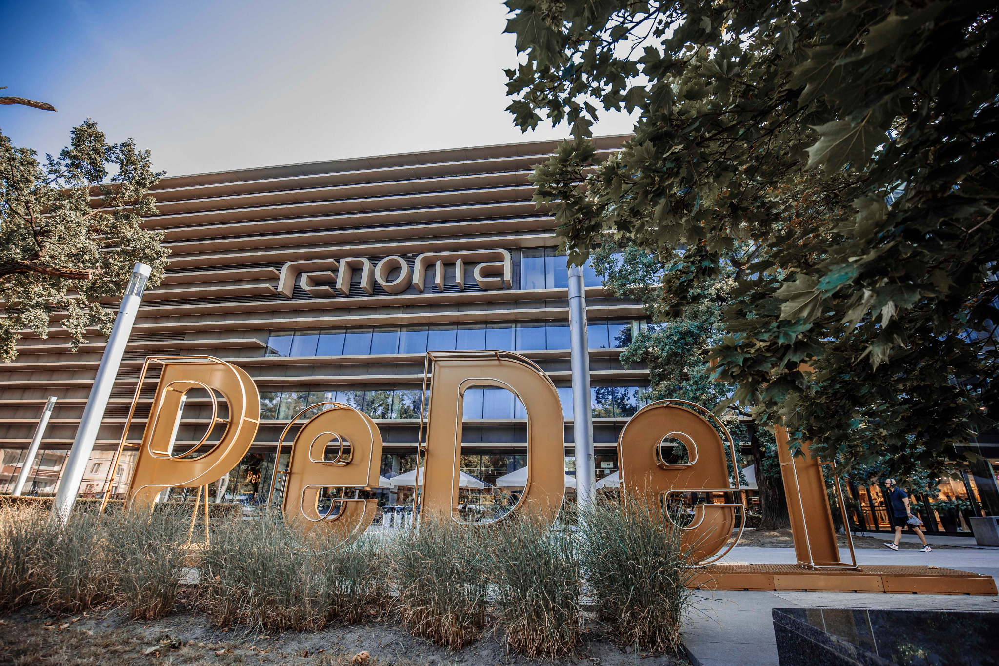 Duże, metalowe litery tworzące nazwę P&O ustawione przed nowoczesnym budynkiem biurowym z przeszkloną fasadą i poziomymi pasami, widoczne trawy ozdobne i drzewa