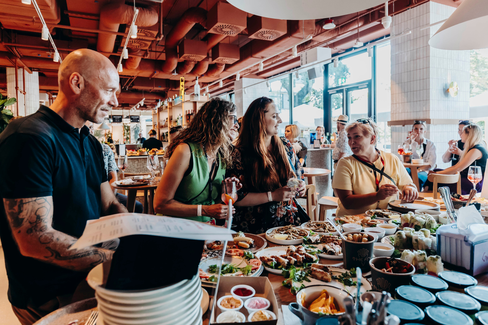 Uczestnicy eventu firmowego przy stole z przekąskami i napojami, widoczne osoby rozmawiające i jedzące, jasne wnętrze z industrialnymi elementami.