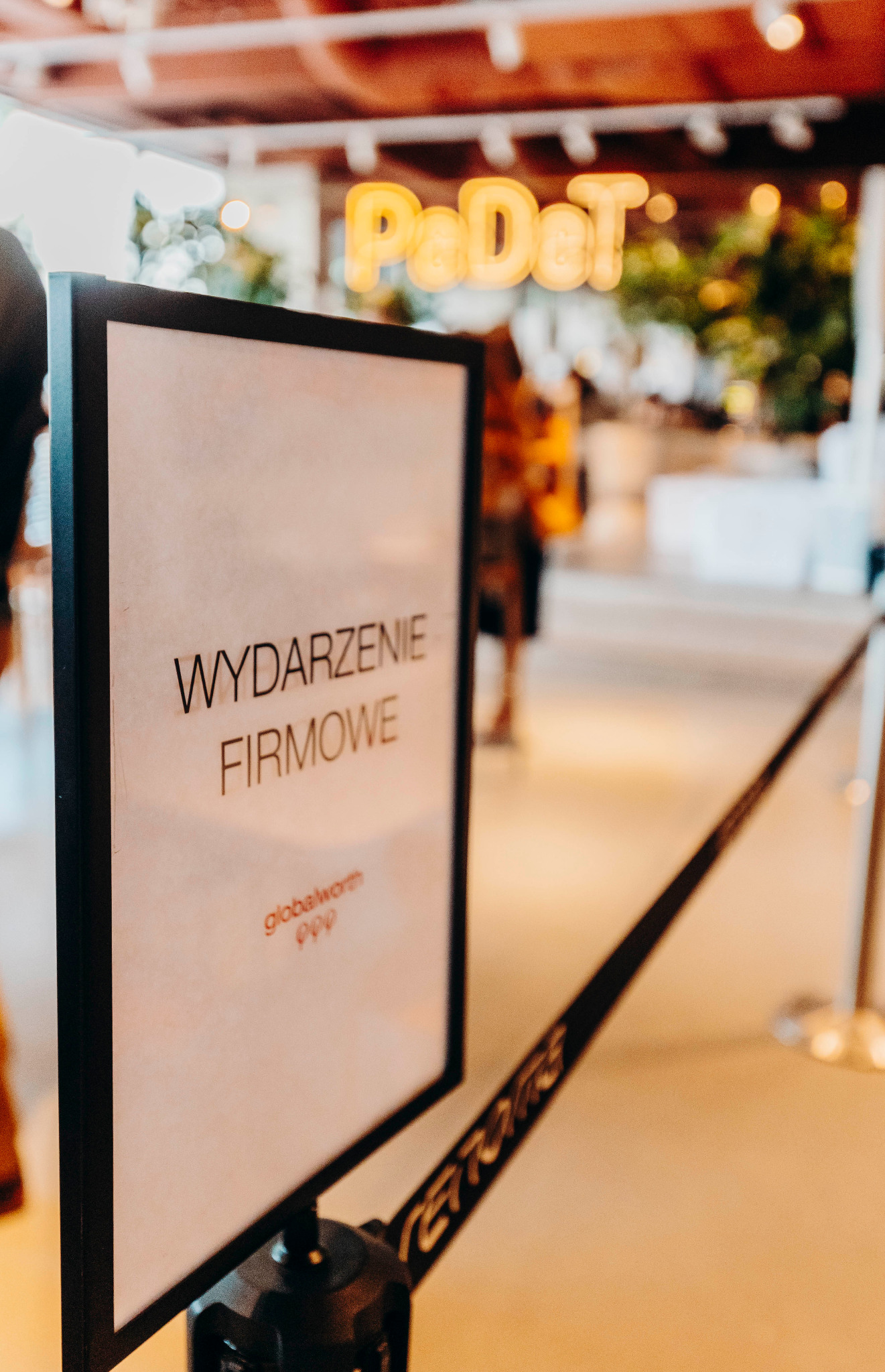Tablica informacyjna z napisem 'WYDARZENIE FIRMOWE' przed wejściem do sklepu, w tle neonowy napis 'PaDdT' i fragment kolejki z taśmą.