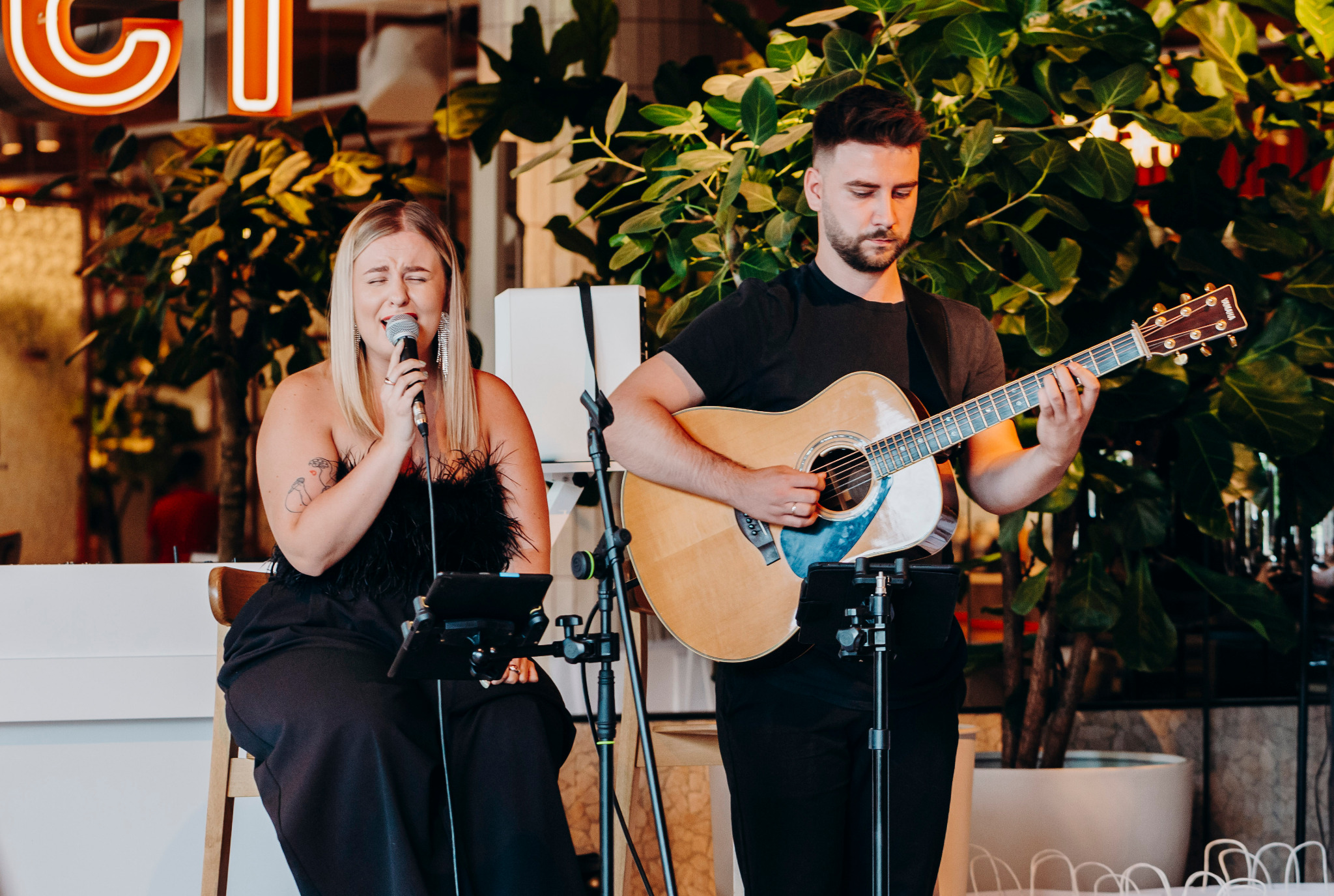 Duet muzyczny: wokalistka z mikrofonem i gitarzysta z gitarą akustyczną występują na tle zielonych roślin i neonowego napisu w lokalu.