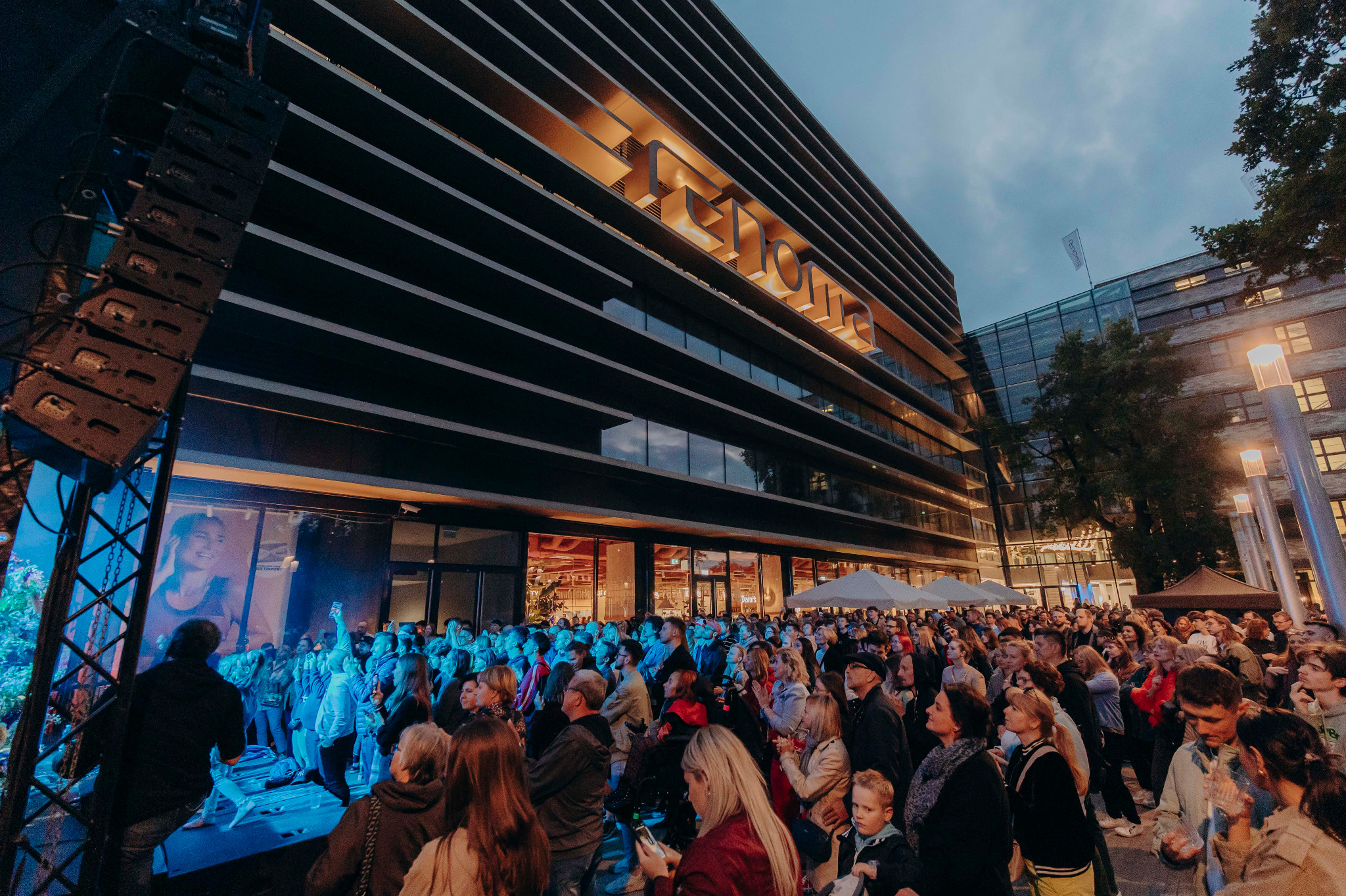 Tłum ludzi zgromadzony przed budynkiem z podświetlonym napisem na fasadzie podczas wydarzenia plenerowego, widoczna scena z oświetleniem i nagłośnieniem.