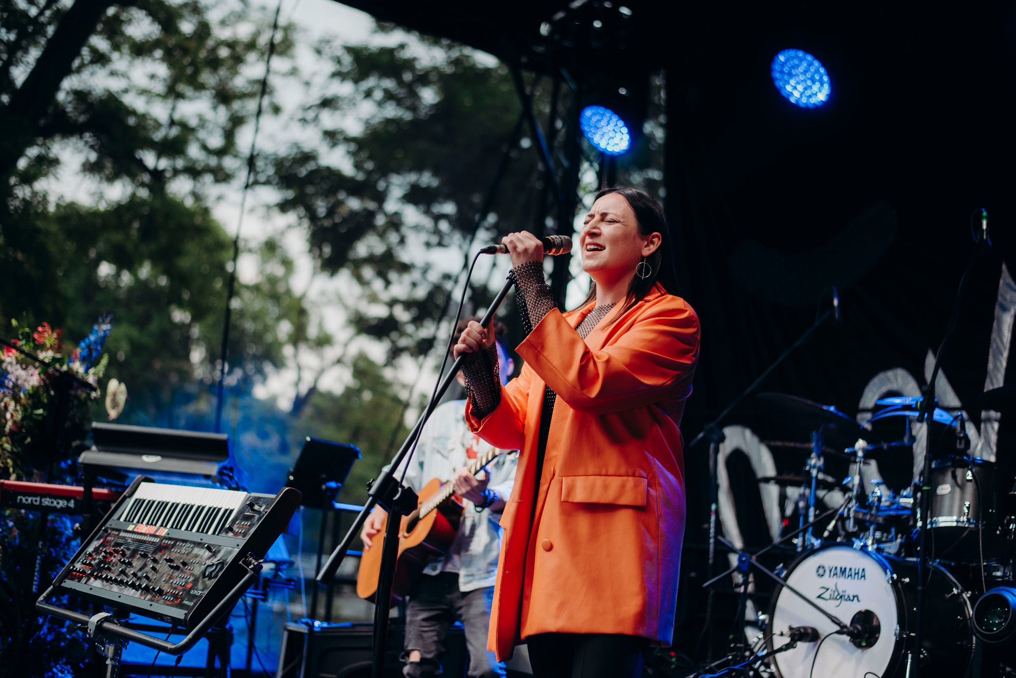 Wokalistka w pomarańczowej marynarce śpiewa do mikrofonu na scenie podczas koncertu, widoczny keyboard i perkusja Yamaha Zildjian.