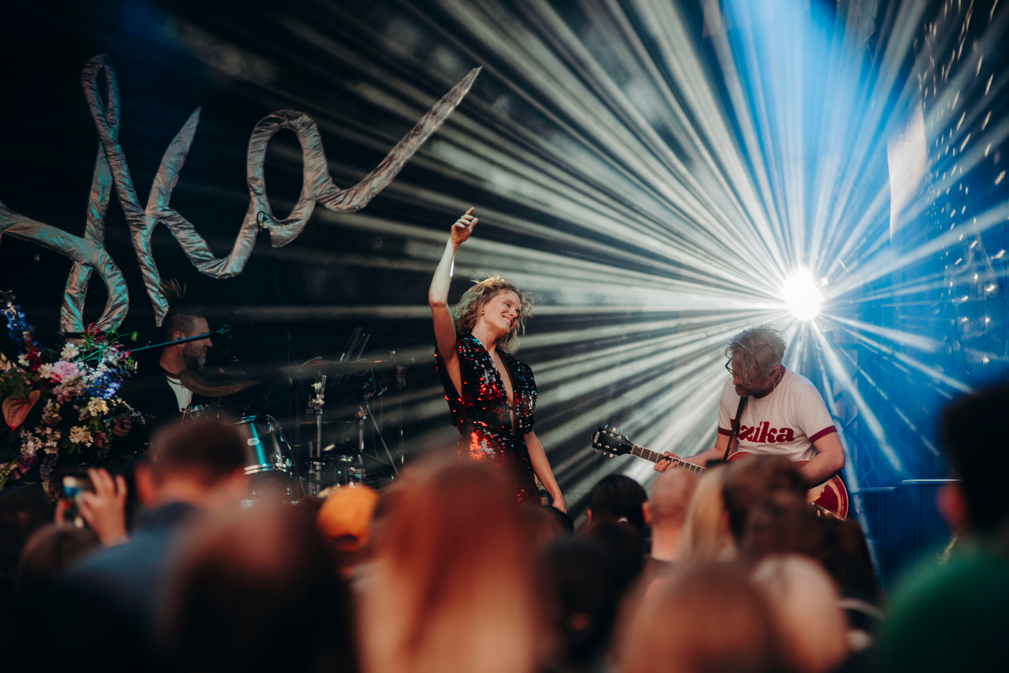 Wokalistka w błyszczącej sukience na scenie podczas koncertu, gitarzysta i perkusista w tle, snopy światła i rozmazany tłum.