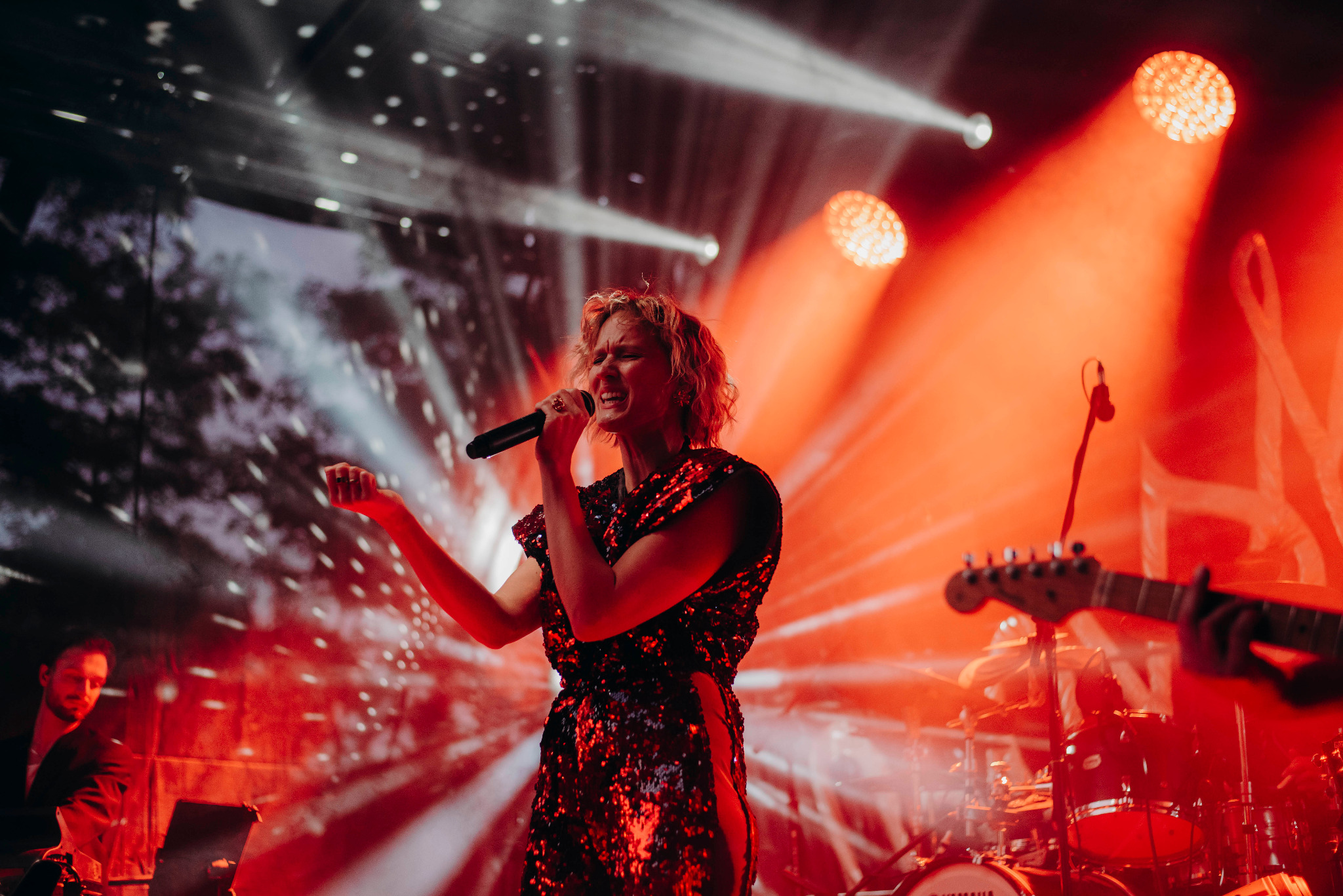 Wokalistka w cekinowej sukience śpiewa na scenie oświetlonej czerwonymi reflektorami, w tle widoczni muzycy i instrumenty.