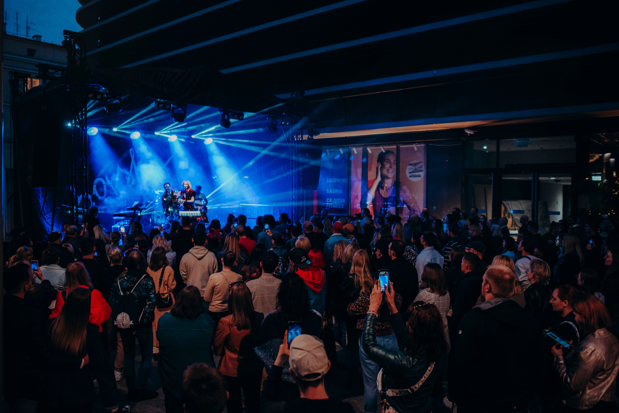 Koncert plenerowy z zespołem na scenie oświetlonej niebieskimi reflektorami, tłum ludzi nagrywających występ telefonami, w tle budynek z podświetlonym banerem reklamowym.