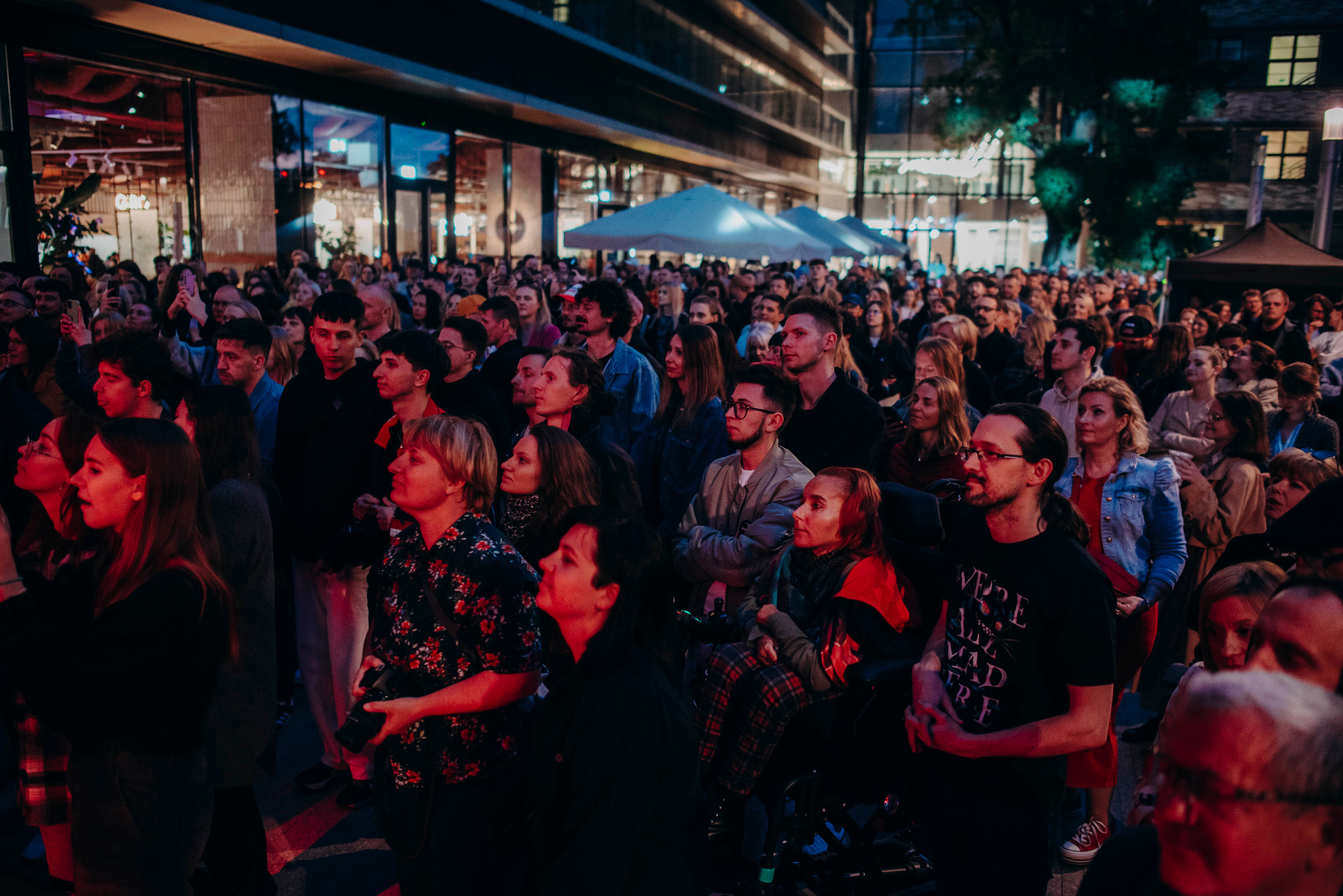 Tłum ludzi zgromadzony na otwartym wydarzeniu plenerowym, oświetlonym wieczornym światłem, z widocznymi białymi namiotami w tle i budynkami w tle.