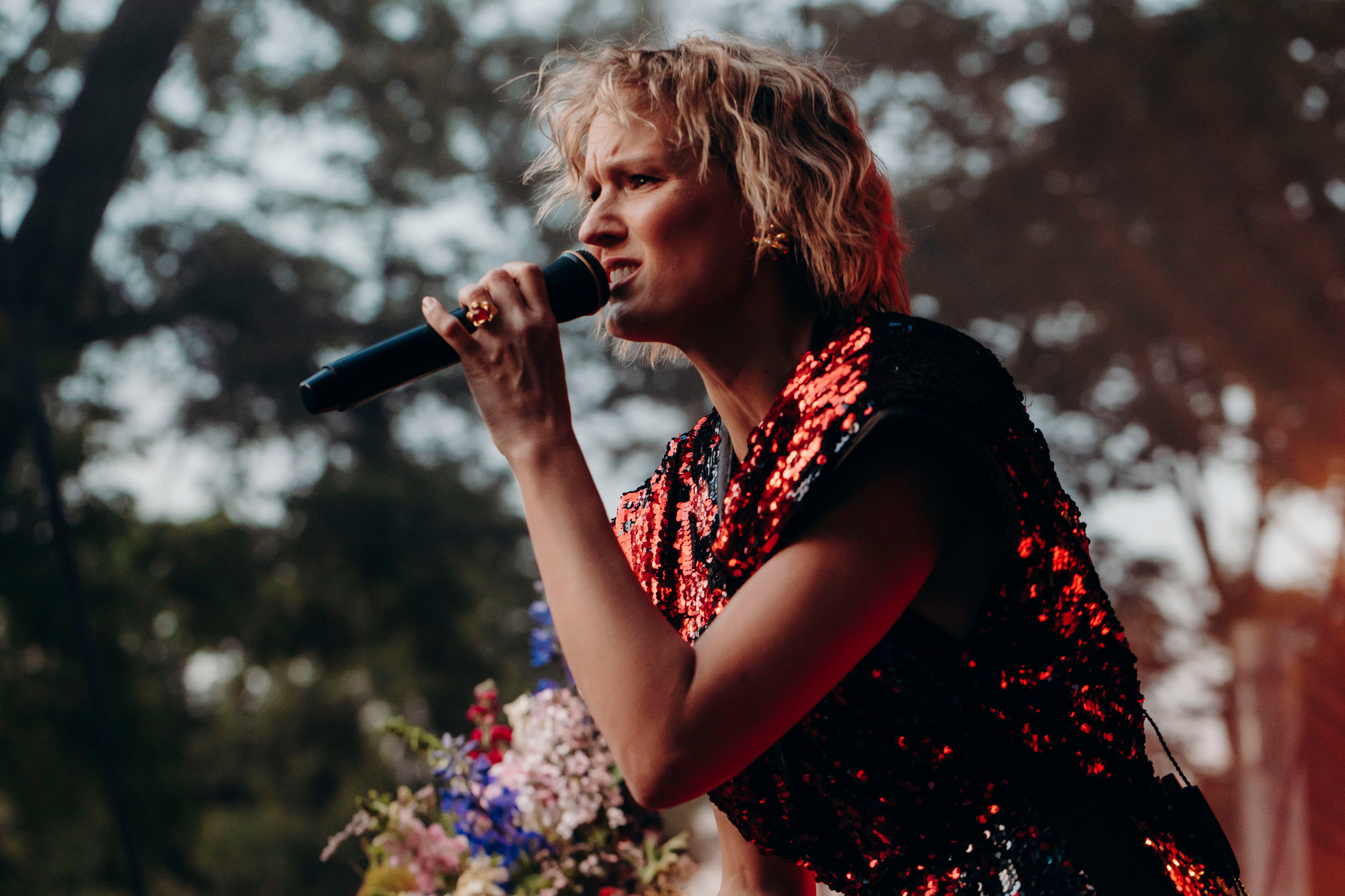 Wokalistka w cekinowej sukience śpiewa do mikrofonu na tle rozmytych drzew i bukietu kwiatów.