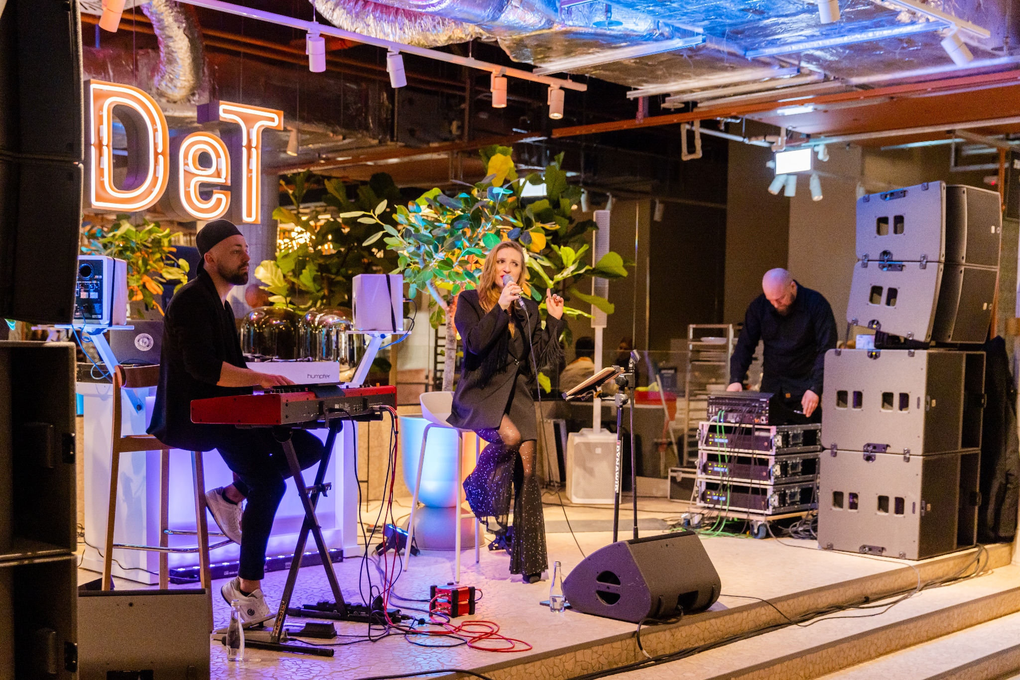 Występ na scenie: wokalistka w czarnym stroju z błyszczącymi nogawkami śpiewa do mikrofonu, akompaniuje jej klawiszowiec na czerwonym keyboardzie, a technik dźwięku obsługuje sprzęt nagłośnieniowy.