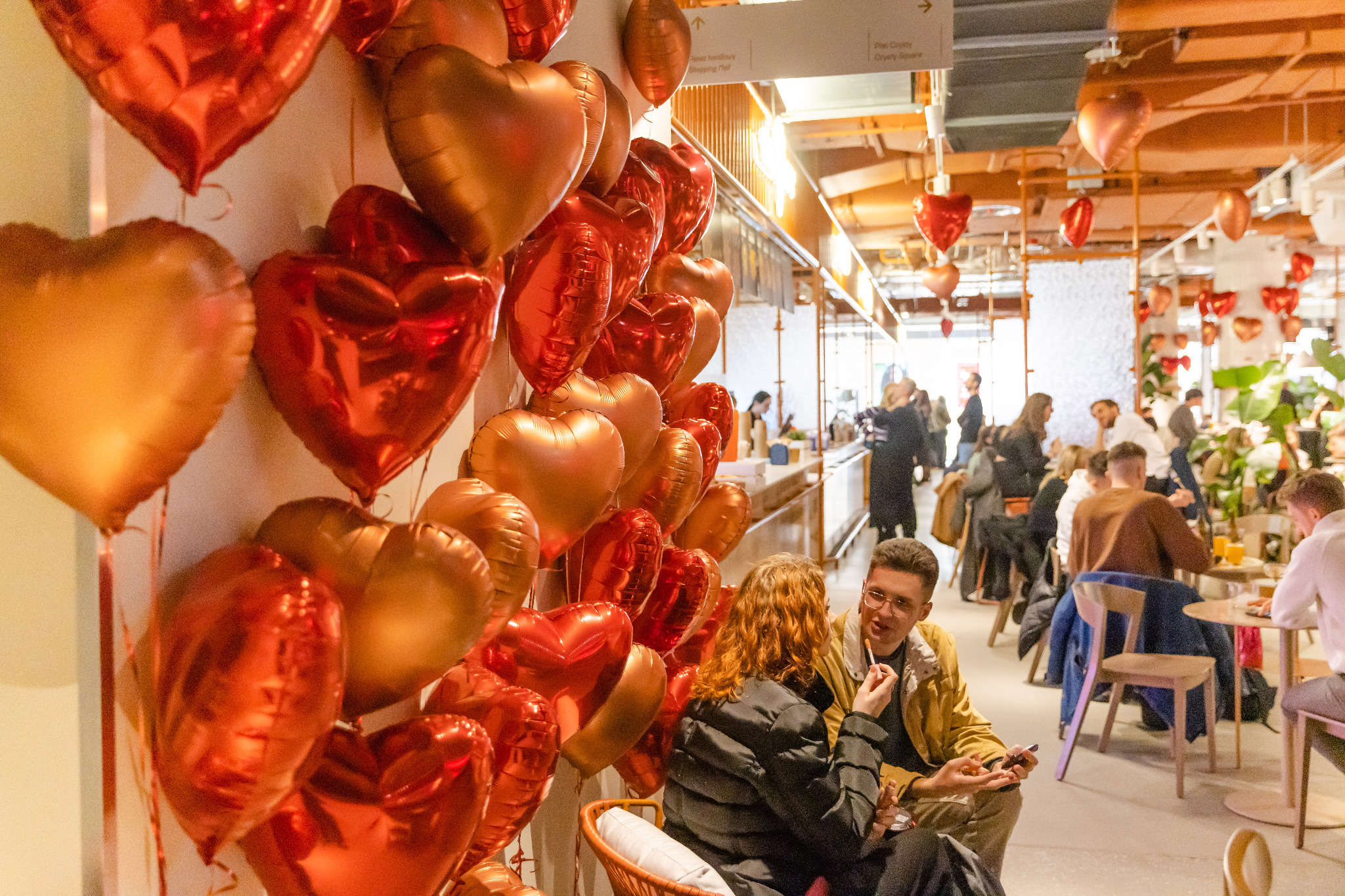 Para siedząca w kawiarni udekorowanej czerwonymi i złotymi balonami w kształcie serc, w tle widoczni inni goście i rośliny doniczkowe.