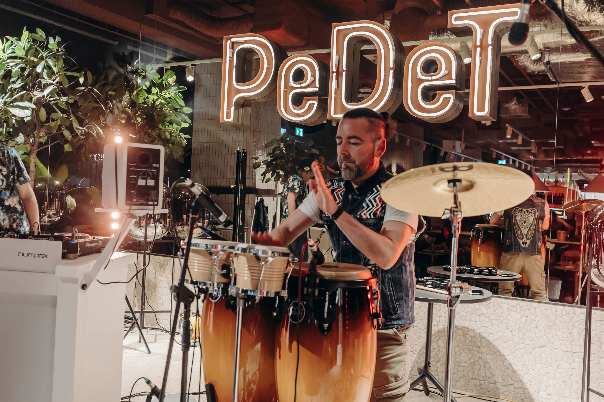 Muzyk grający na bębnach conga podczas wydarzenia, w tle neonowy napis 'PeDeT', rośliny doniczkowe i sprzęt DJ-ski.
