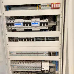 Wnętrze rozdzielnicy elektrycznej z zainstalowanymi licznikami energii, zabezpieczeniami nadprądowymi i ogranicznikami przepięć, widoczne okablowanie i listwy zaciskowe.