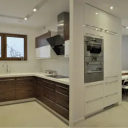 Nowoczesna kuchnia z drewnianymi szafkami dolnymi, białymi górnymi, i wbudowanym sprzętem AGD ze stali nierdzewnej, oświetlona punktowymi lampami sufitowymi.