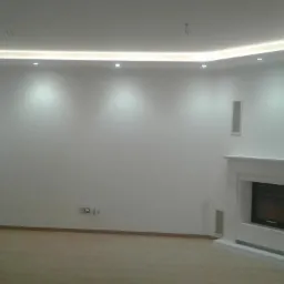 Białe wnętrze pokoju z kominkiem, oświetleniem LED na suficie i panelami podłogowymi, widok po remoncie.