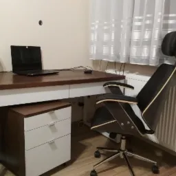 Drewniany blat biurka z białymi nogami i szafką z szufladami, nowoczesne krzesło biurowe z wysokim oparciem, laptop i lampka na biurku, widok na pokój z zasłonami i grzejnikiem.