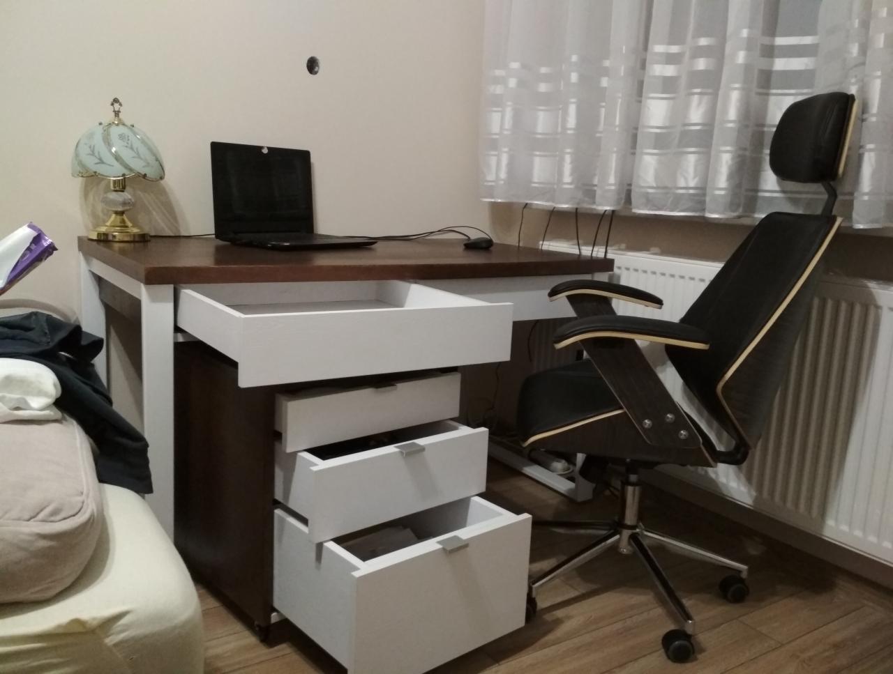 Biurko z ciemnym drewnianym blatem i białymi szufladami, z otwartą górną szufladą, obok stoi czarne krzesło biurowe z drewnianymi elementami, na blacie laptop i lampka.
