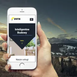 Dłoń trzymająca smartfon z otwartą stroną internetową firmy budowlanej USTR na tle górskiego krajobrazu.