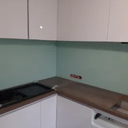 Montaż szklanego panelu ściennego w kuchni, widoczne białe szafki, fragment płyty grzewczej i niezabezpieczone kable elektryczne w ścianie.