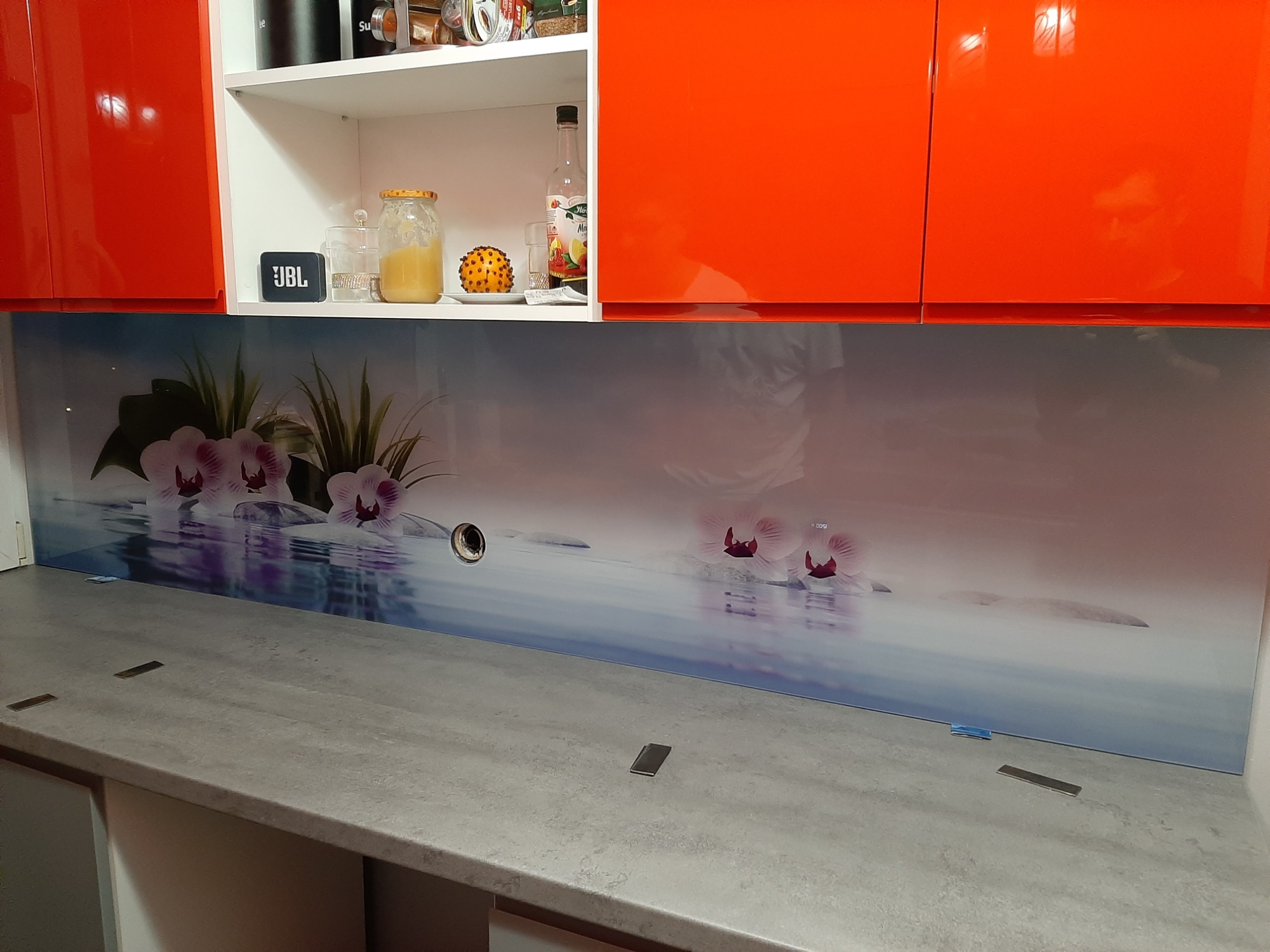 Szklany panel kuchenny z motywem kwiatów orchidei, zamontowany między blatem a szafkami, widoczne otwory na gniazdka elektryczne.