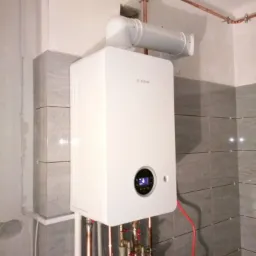 Nowoczesny, biały piec gazowy marki Bosch zamontowany na tle ściany wyłożonej szarymi płytkami, z widocznymi miedzianymi rurami instalacji i podłączeniami.