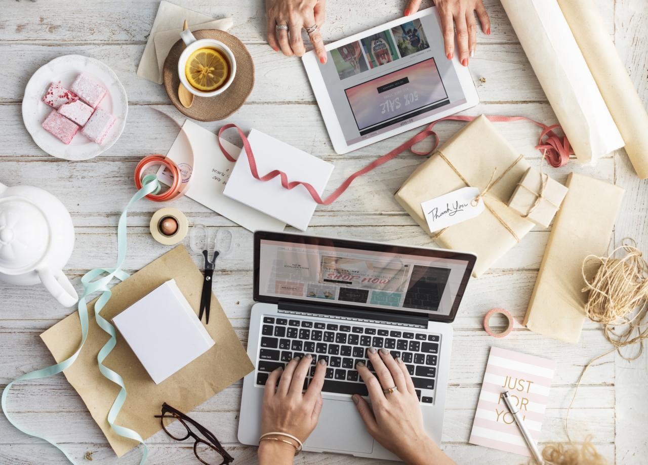 Widok z góry na biurko z laptopem, tabletem, zapakowanymi prezentami, wstążkami, herbatą z cytryną i słodyczami, sugerujący przygotowywanie spersonalizowanych upominków.