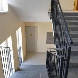 Klatka schodowa w budynku wielorodzinnym po odświeżeniu, widoczne szare płytki na schodach i podłodze, kremowe ściany oraz czarne metalowe balustrady.