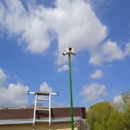 Trzy kamery monitoringu na zielonym słupie, widoczne z dołu na tle błękitnego nieba z białymi chmurami, obok drabina aluminiowa oparta o budynek z garażami.