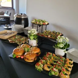 Stół cateringowy z burgerami, wrapami, sałatkami i kanapeczkami na czarnym obrusie, dodatkowo białe kwiaty w doniczkach jako dekoracja.