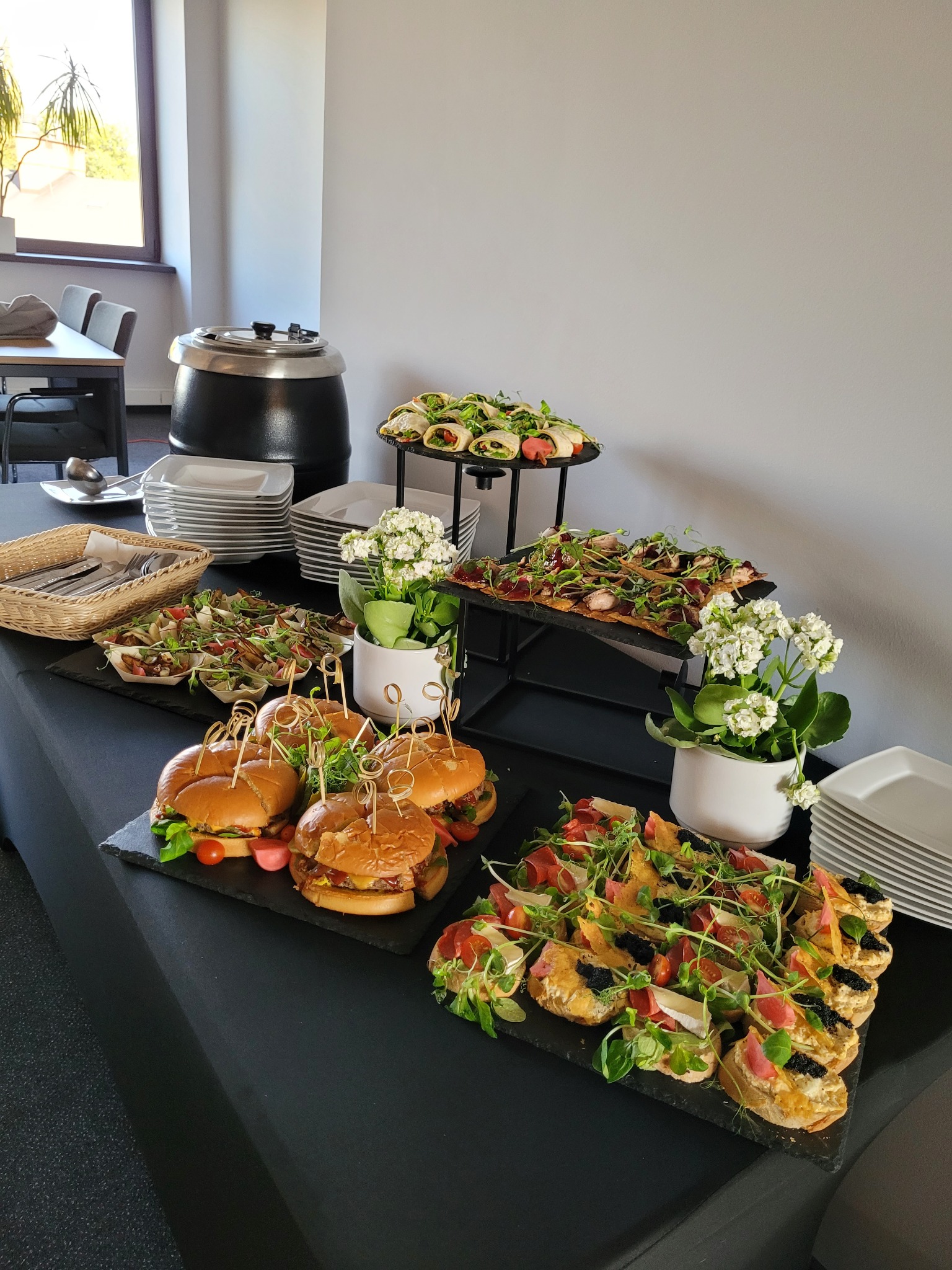 Stół cateringowy z burgerami, wrapami, sałatkami i kanapeczkami na czarnym obrusie, dodatkowo białe kwiaty w doniczkach jako dekoracja.