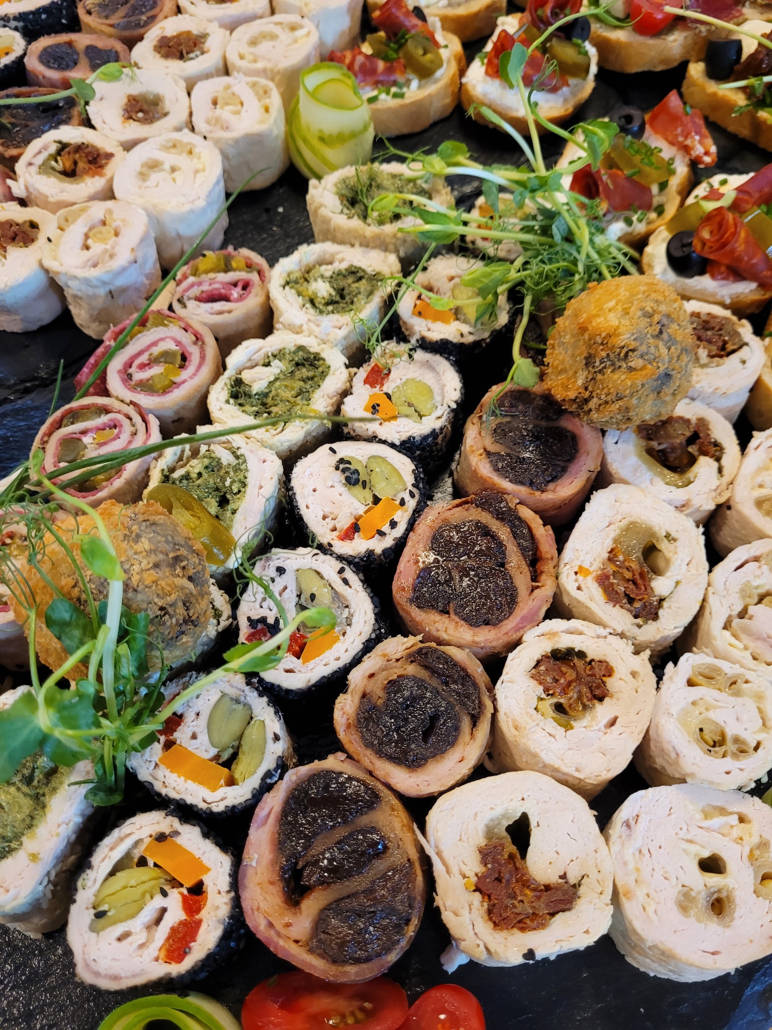 Dekoracyjnie ułożone, różnorodne roladki i kanapeczki na ciemnej desce, z widocznymi detalami nadzienia i posypki, udekorowane świeżymi pędami.