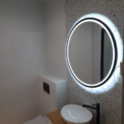 Nowoczesna łazienka z owalną umywalką nablatową, czarną armaturą i okrągłym lustrem podświetlanym LED, na tle ściany wykończonej mozaiką.