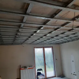 Stelaż sufitowy z profili metalowych w trakcie montażu, widoczne przewody, okno z widokiem na zieleń, grzejnik i narzędzia na podłodze w surowym pomieszczeniu.