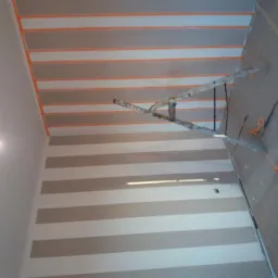 Ściana w trakcie malowania w poziome pasy, zabezpieczona taśmą malarską, z widoczną drabiną, kuwetami malarskimi i resztkami taśmy na podłodze.